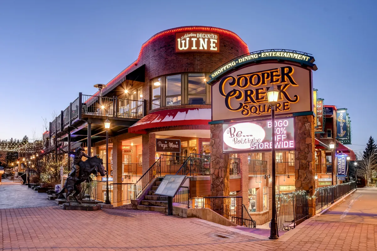 A welcoming street-level view of Cooper Creek Square, featuring the iconic sign, a wine shop, outdoor seating, and a horse statue. The area is full of life, hinting at ongoing live music, seasonal festivals, shopping, and group-friendly dining.