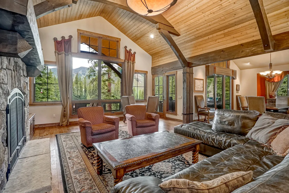 A beautifully designed living area with large windows offering panoramic mountain views. The space includes a substantial stone fireplace, leather sofas, exposed wooden beams, and an area rug, evoking a cozy yet elegant atmosphere perfect for relaxing after a day of adventures.