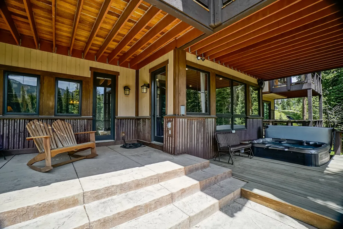 A cozy wooden deck featuring a bubbling hot tub, Adirondack chairs, and expansive windows that glow warmly from within. Snow gently dusts the surroundings, and the scene transitions to evening, encouraging relaxation after a day of adventure.