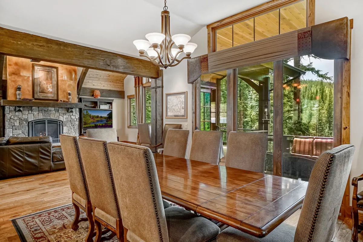 A cozy dining area with a substantial wooden table beneath a chandelier, adjacent to large windows and a stone fireplace. The atmosphere is perfect for family dinners, celebrations, or intimate gatherings, with natural light and warmth enhancing every meal.
