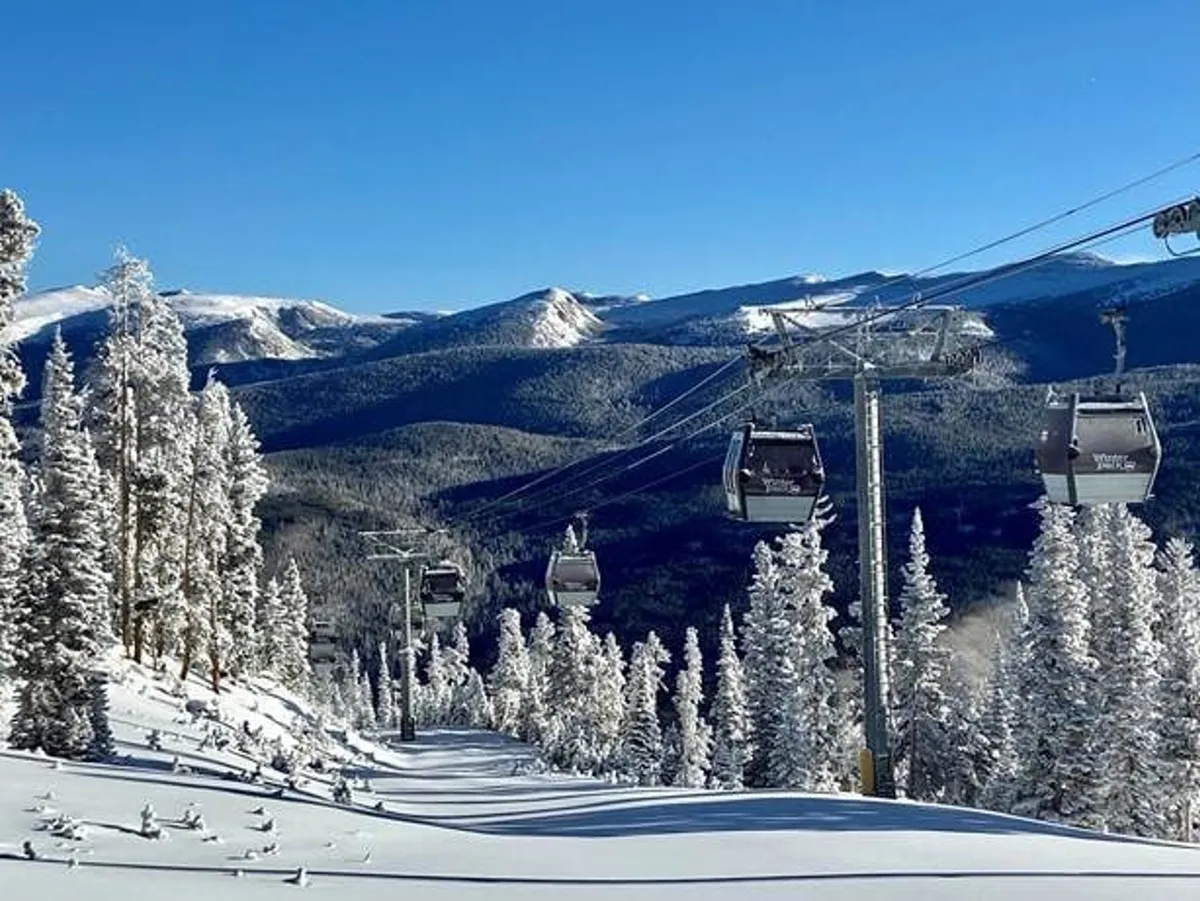A panoramic view of a ski lift rising over snow-covered trees and a majestic mountain range. The landscape is blanketed in fresh white snow, creating a classic alpine winter scene. Skiers and snowboarders can be seen gliding down the slopes, while the crisp air captures the essence of adventure and excitement.
