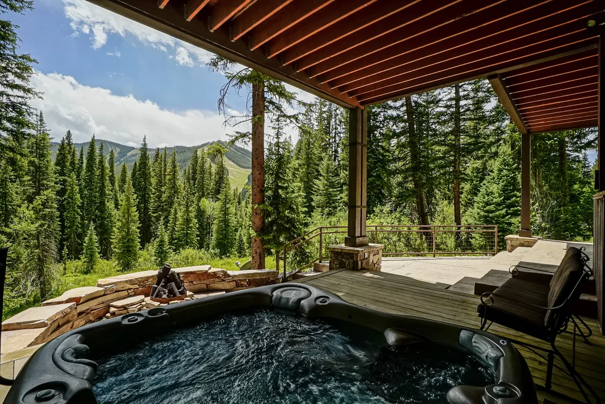 This exterior scene showcases a tranquil wooden deck surrounded by pine trees and sweeping mountain views. A bubbling hot tub is the focal point, offering a relaxing spot under open skies. Adirondack chairs provide comfortable seating, inviting guests to soak, chat, or enjoy the scenery.