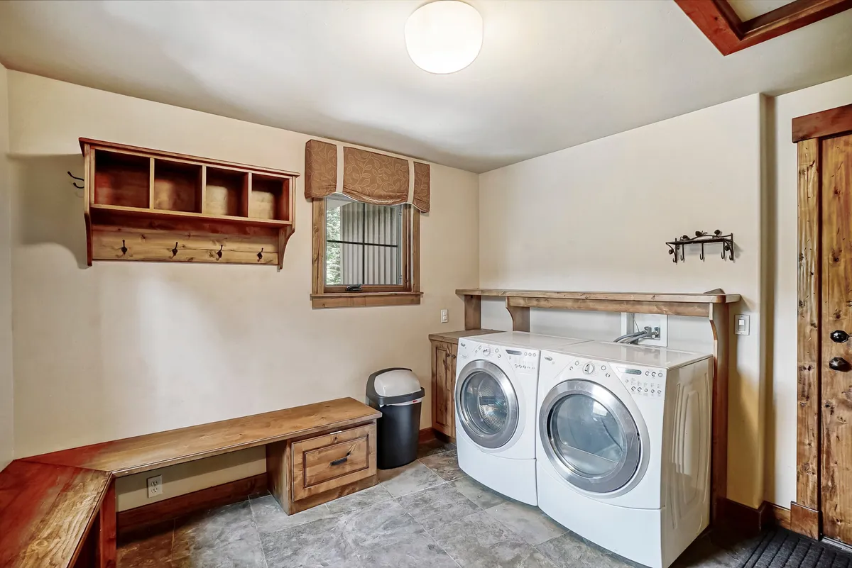 This image shows a practical and welcoming laundry room featuring a washer and dryer, a wooden bench for convenience, a shelving unit for organization, and a window with a decorative valance letting in natural light. The scene is clean and orderly, emphasizing the utility and warmth of the space.
