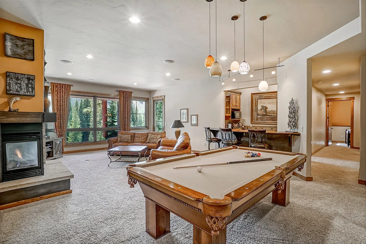 An inviting interior great room featuring a full-sized pool table as the focal point, complemented by a cozy stone fireplace, large windows offering ample natural light, and luxurious leather seating arranged for both gaming and relaxation. The scene is set for socializing, unwinding after outdoor adventures, or hosting lively family tournaments.