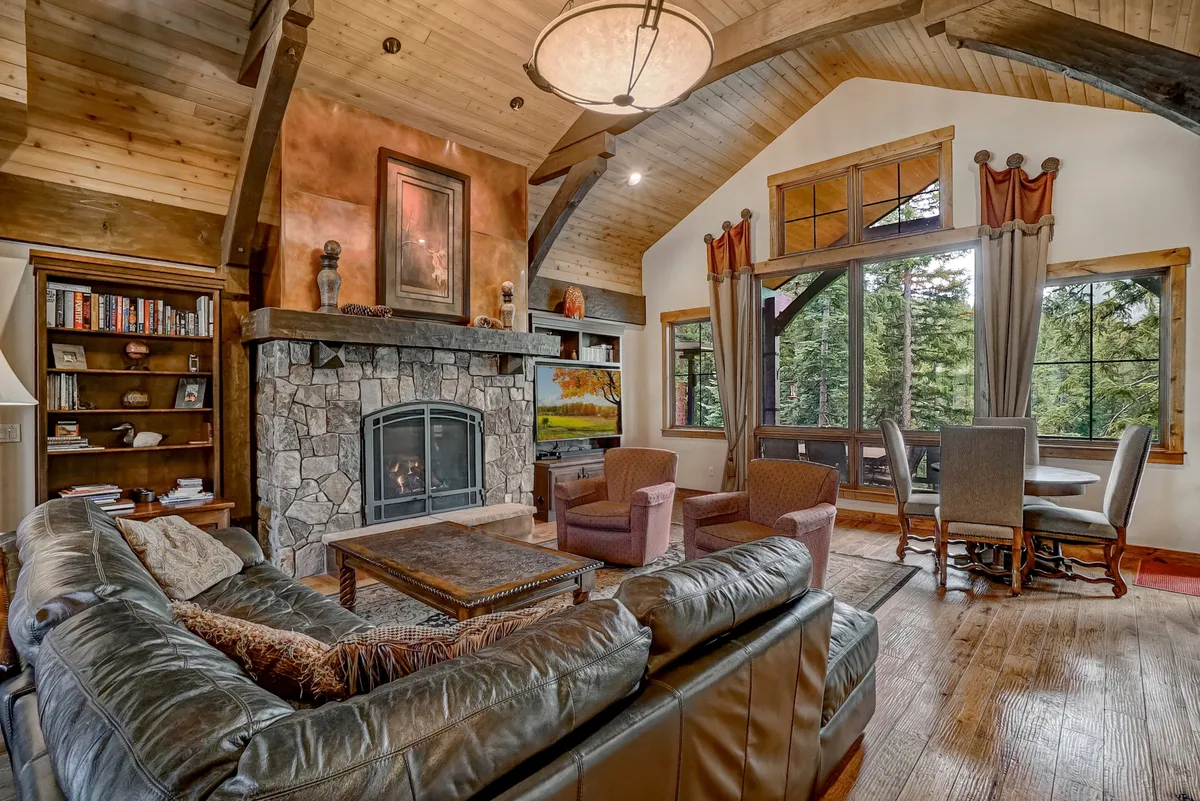 A spacious great room features a stone fireplace as the focal point, with large floor-to-ceiling windows letting in ample mountain light. Comfortable seating is arranged around the fireplace and a bookshelf, and an adjacent dining area is visible. The room exudes warmth and is clearly designed for gatherings, relaxation, and shared moments.