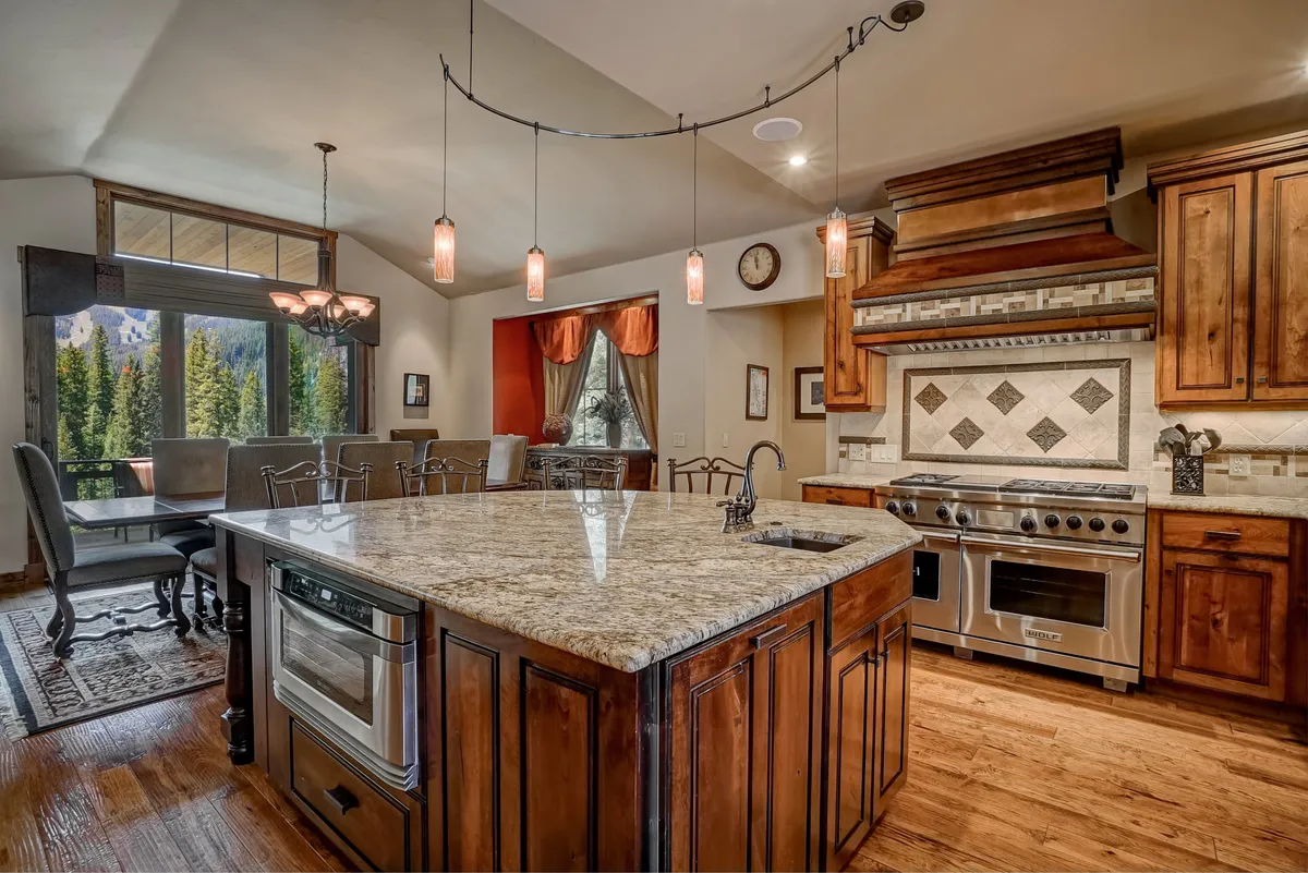 An inviting interior shot of a spacious, modern kitchen with a granite island, high-end appliances, warm wooden cabinetry, and a large window showcasing natural light. The space is set up for group cooking, casual conversations, and meal gatherings.