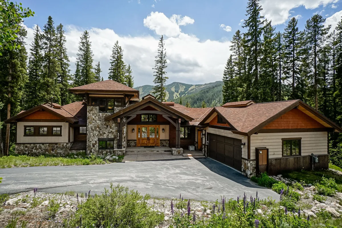 The image showcases the impressive stone facade and wooden doors of a luxury mountain home, framed by lush surrounding trees and set against a breathtaking mountain backdrop. Large windows promise sweeping views from inside, while the welcoming porch invites guests to unwind and take in spectacular sunsets. The serene setting highlights the property's blend of rustic elegance and modern comfort.
