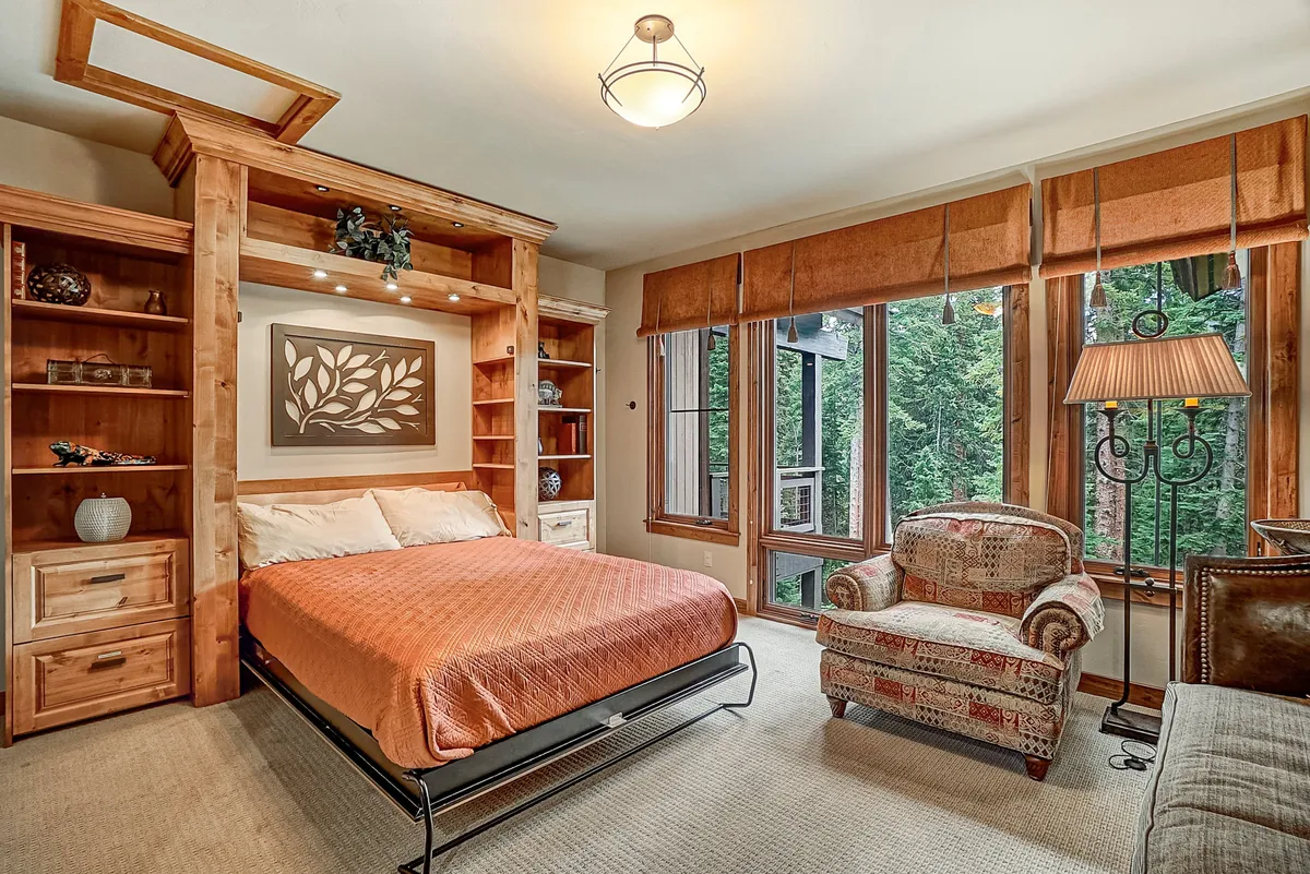 A serene bedroom featuring a comfortable bed, large windows that let in abundant natural light, an inviting armchair, and built-in bookshelves. The room is styled with tranquil colors and offers forested views outside, embodying a peaceful retreat ideal for relaxation.