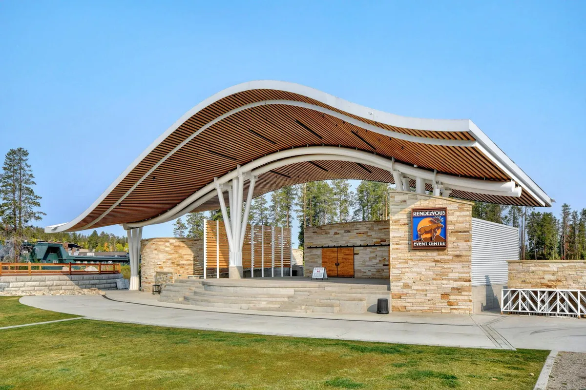An exterior view of a modern structure with a curved wooden roof and stone walls, featuring an open stage area. The setting is ideal for outdoor concerts, community events, and seasonal celebrations, with natural surroundings hinting at the mountain environment.