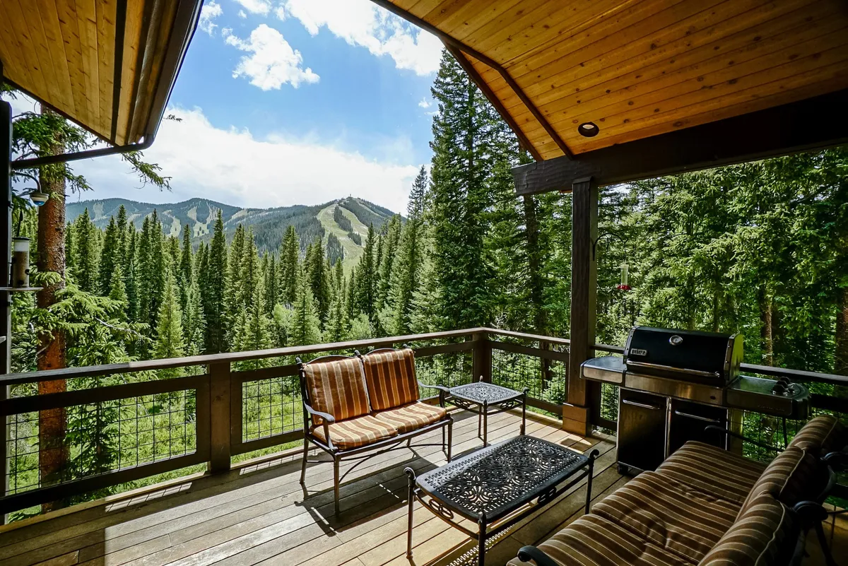 The exterior wooden deck features comfortable seating, a grill, and panoramic mountain views with lush forest surroundings. The atmosphere is relaxed, inviting guests to unwind after a day outdoors.