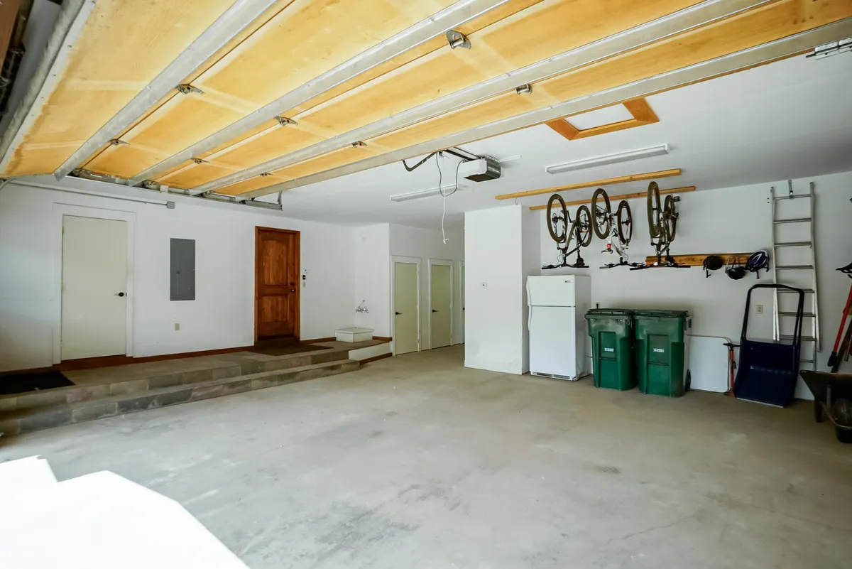 The image depicts an organized garage area with bicycles mounted on the wall, a fridge for extra storage, garbage bins arranged neatly, and a stairway entrance for easy access to the home. The space is clean and ready for storing sports equipment or prepping for outdoor excursions.