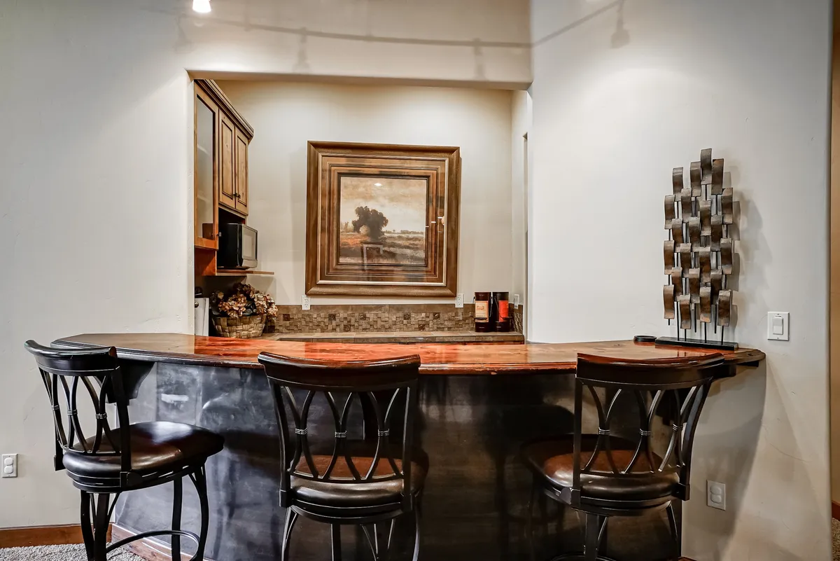A stylish wet bar setup inside, featuring a sleek bar counter with granite finish, bar stools for seating, and framed artwork adorning the walls. The ambiance invites socializing over drinks and snacks, making it an ideal support zone for gatherings in the adjacent recreation area.