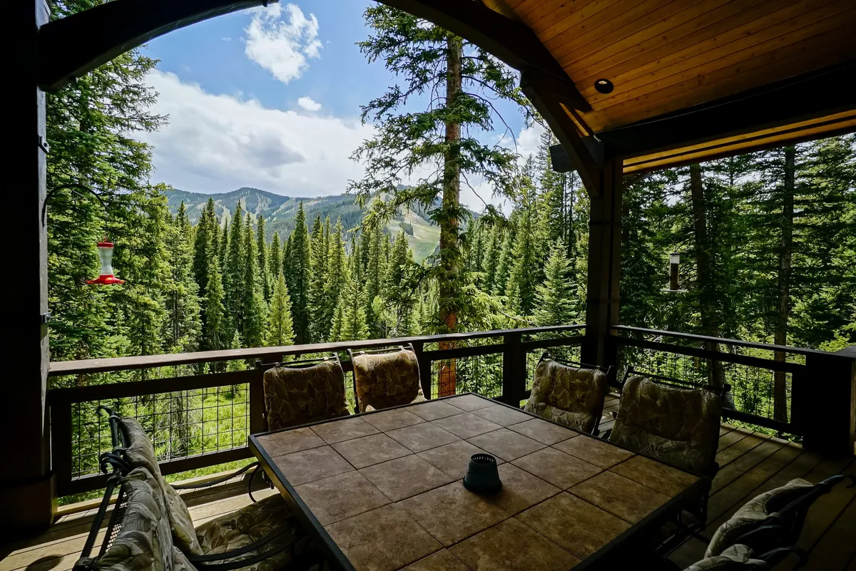 A spacious wooden deck furnished with outdoor chairs and a table, surrounded by tall trees and offering a sweeping mountain view. The deck invites guests to relax, enjoy a meal, or gaze quietly at the wilderness. The tranquil natural setting hints at opportunities for wildlife watching and peaceful moments outdoors.